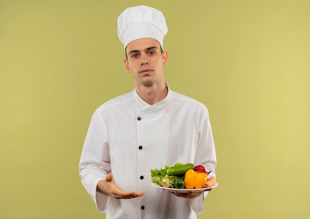 Jeune homme cuisinier portant l'uniforme de chef montrant des légumes sur une assiette à la main sur un mur vert isolé avec copie espace