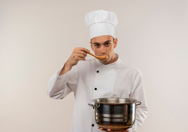 Jeune homme cuisinier portant l'uniforme de chef tenant une casserole en essayant de soupe de cuillère sur mur blanc isolé