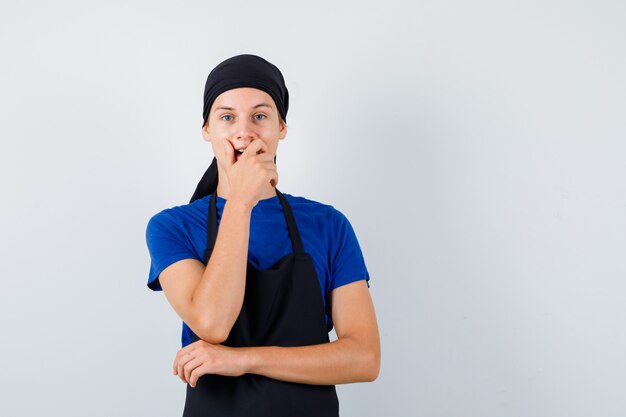 Jeune homme cuisinier en t-shirt, tablier avec la main sur la bouche et l'air étonné, vue de face.