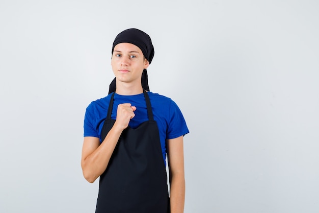 Jeune homme cuisinier en t-shirt, tablier avec poing fermé sur la poitrine et l'air confiant, vue de face.