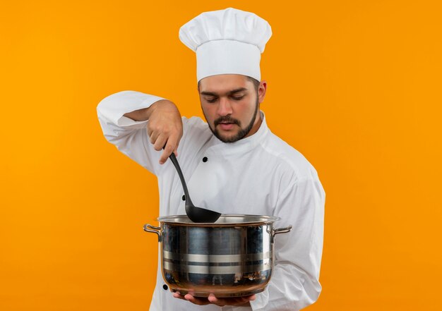 Jeune homme cuisinier en uniforme de chef tenant la louche et le pot à l'intérieur du pot isolé sur l'espace orange