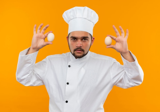 Photo gratuite jeune homme cuisinier en uniforme de chef tenant des oeufs et à la recherche d'isolement sur l'espace orange