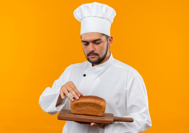 Jeune homme cuisinier en uniforme de chef tenant une planche à découper avec du pain dessus et toucher du pain isolé sur l'espace orange