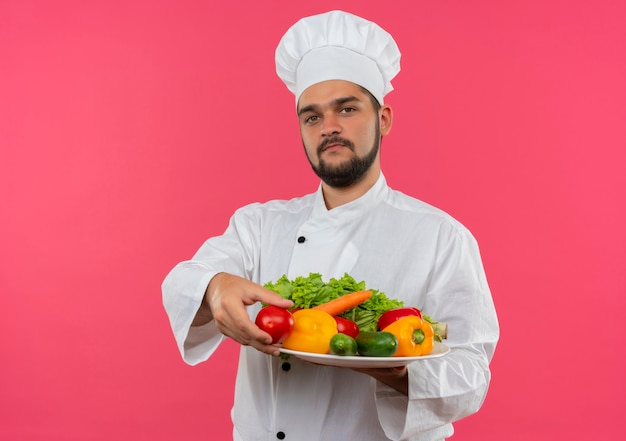 Jeune homme cuisinier en uniforme de chef tenant la plaque de légumes et de tomates et à la recherche d'isolement sur l'espace rose