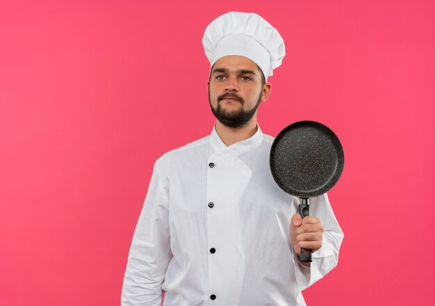 Jeune homme cuisinier en uniforme de chef tenant une poêle et regardant tout droit isolé sur l'espace rose