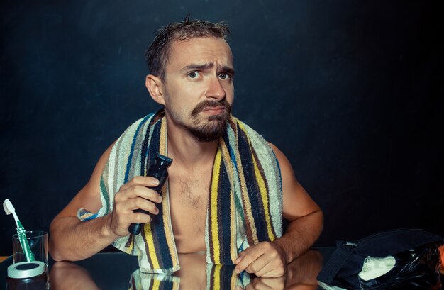 Le jeune homme dans la chambre assis devant le miroir se gratte la barbe à la maison. Concept d'émotions humaines et de style de vie