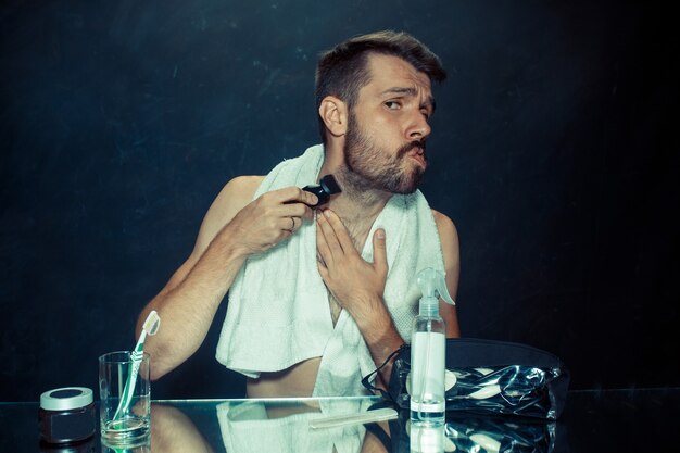 Le jeune homme dans la chambre assis devant le miroir se gratte la barbe à la maison. Concept d'émotions humaines