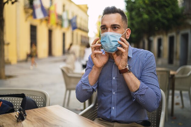 Jeune homme dans une chemise bleue portant un masque médical assis dans un café en plein air - concept Covid-19