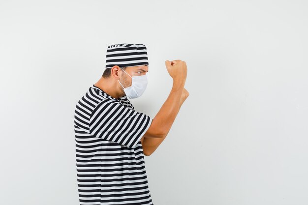 Jeune homme debout en boxeur pose en t-shirt rayé, chapeau, masque et à la recherche résolue.