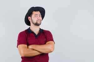 Photo gratuite jeune homme debout avec les bras croisés en t-shirt, chapeau et à la satisfaction de soi. vue de face.