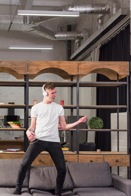 Jeune homme debout sur le canapé avec un casque sur sa tête danser à la maison