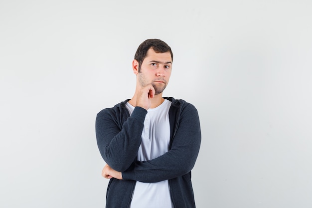 Jeune homme debout dans la pensée pose en t-shirt blanc et sweat à capuche noir zippé sur le devant et à la vue de face sérieuse