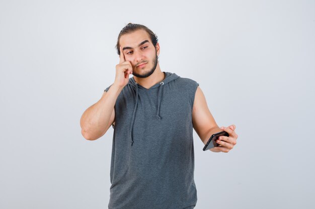 Jeune homme debout dans la pensée pose, tenant le téléphone à la main en t-shirt à capuche et à la vue sensible, de face.