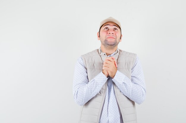Jeune homme debout dans la pose de prière et regardant vers le haut en veste beige et casquette et à la recherche concentrée. vue de face.