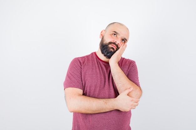 Jeune homme debout dans une pose de réflexion tout en s'appuyant sur la joue sur la paume en t-shirt rose et l'air pensif, vue de face.