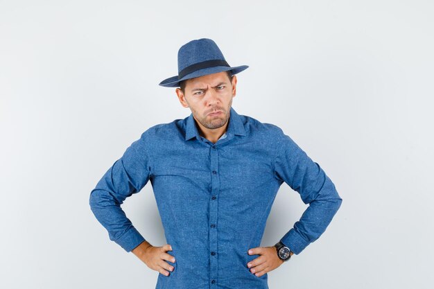 Jeune homme debout avec les mains sur la taille en chemise bleue, chapeau et l'air nerveux, vue de face.