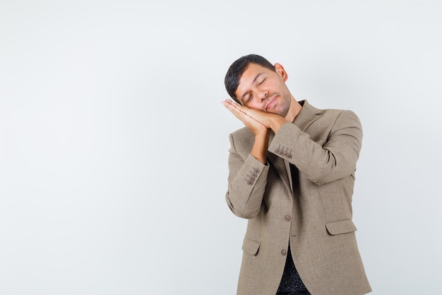 Jeune homme debout tout en faisant un geste d'oreiller en veste marron grisâtre et ayant l'air endormi, vue de face. espace libre pour votre texte