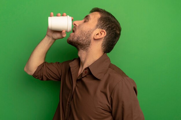 Jeune homme debout en vue de profil, boire du café dans une tasse de café en plastique isolé sur un mur vert