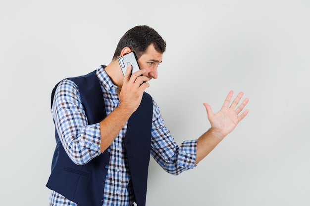 Jeune homme discutant de quelque chose sur téléphone mobile en chemise, gilet, vue de face.