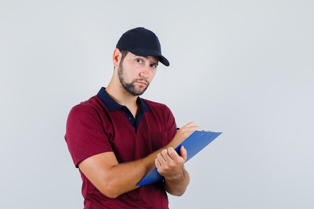 Jeune homme écrivant quelque chose sur son cahier en t-shirt rouge, casquette noire et à la recherche concentrée, vue de face.