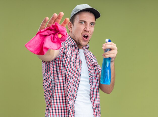 Un jeune homme effrayé portant une casquette tenant un agent de nettoyage avec un chiffon montrant un geste correct isolé sur fond vert olive