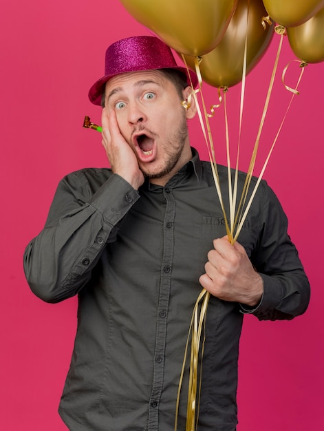 Jeune homme effrayé portant chapeau rose tenant des ballons avec souffleur de fête mettant la main sur la joue isolé sur rose