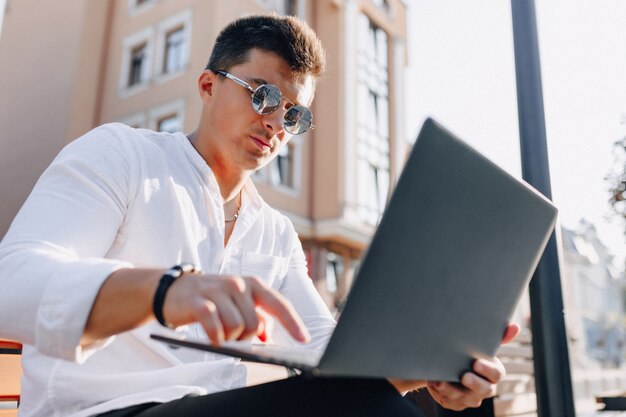 Jeune homme élégant en chemise avec téléphone et ordinateur portable sur un banc par une chaude journée ensoleillée à l'extérieur, freelance