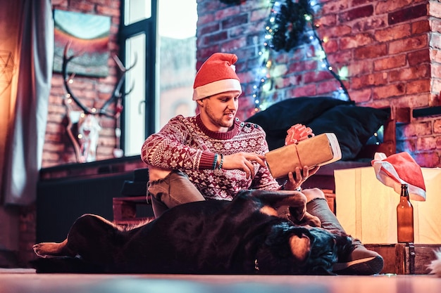 Photo gratuite un jeune homme élégant tenant une boîte-cadeau tout en étant assis avec son mignon chien dans un salon décoré au moment de noël.