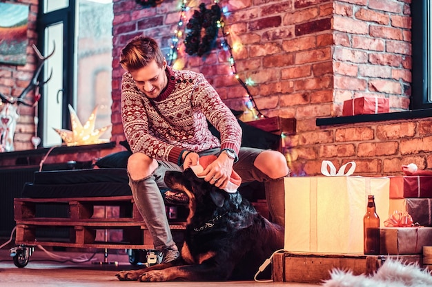 Un jeune homme élégant tenant une boîte-cadeau tout en étant assis avec son mignon chien dans un salon décoré au moment de Noël.