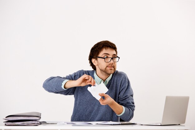 Jeune homme, employé agissant fou au travail, asseoir le bureau, déchirer des documents, se sentir tendu, épuisé au travail, détourner le regard