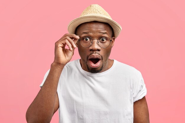 Un jeune homme ému et stupéfait ouvre la bouche et regarde à travers des lunettes, choqué par les dernières nouvelles, porte un chapeau de paille à la mode, se tient seul contre le mur rose