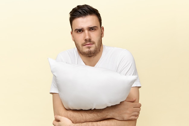 Jeune homme endormi posant avec coussin. Mec attrayant avec des poils ayant une expression faciale fatiguée à cause de l'insomnie essayant de s'endormir, tenant un oreiller.