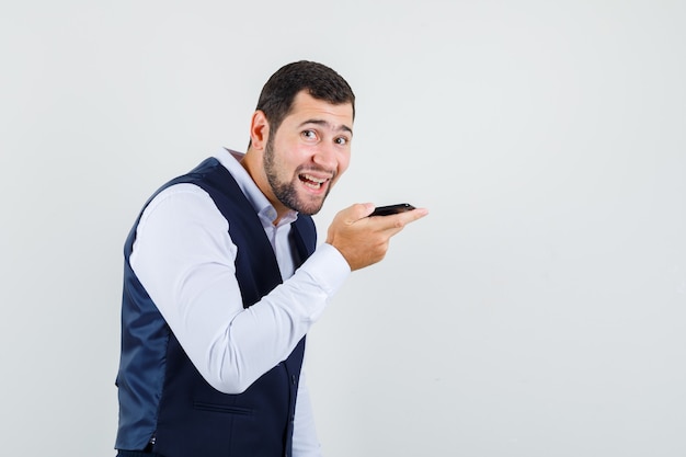 Jeune homme enregistrant un message vocal sur téléphone mobile en costume, gilet