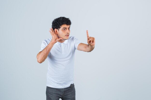 Jeune homme entendant une conversation privée, pointant vers le haut dans un t-shirt blanc, un pantalon et l'air choqué, vue de face.