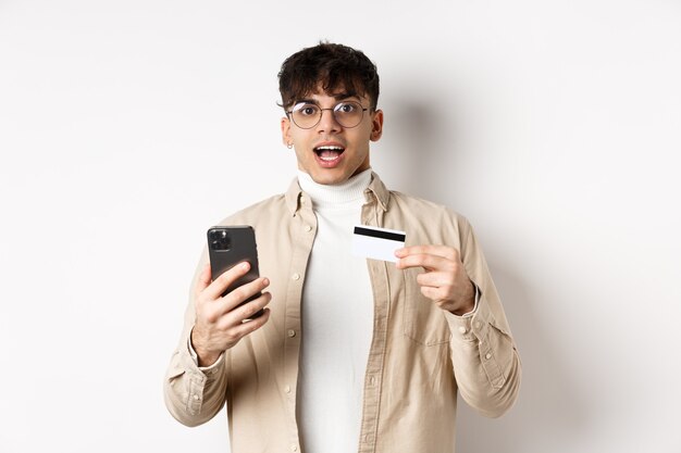 Jeune homme enthousiaste faisant ses achats en ligne, tenant un téléphone portable et une carte de crédit en plastique, effectuant des achats sur Internet, debout sur fond blanc.
