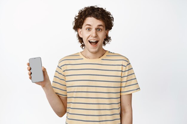 Jeune homme enthousiaste montrant un écran mobile et souriant démontrant son interface d'application fond blanc