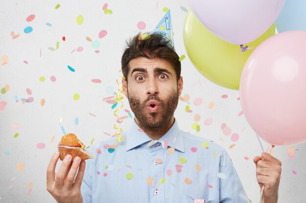 Jeune homme entouré de confettis tenant un petit gâteau et des ballons