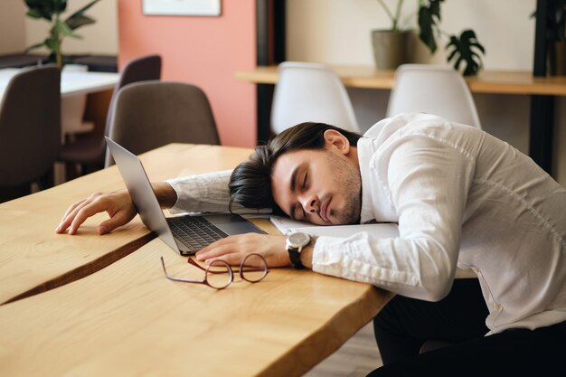 Jeune homme épuisé assis à la table avec un ordinateur portable pendant qu'il dormait au travail dans un bureau moderne