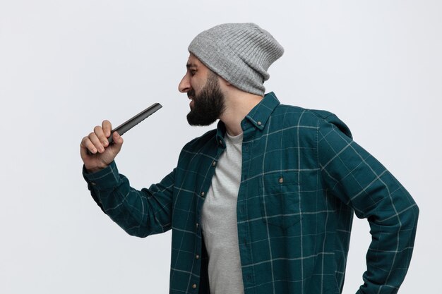 Jeune homme espiègle portant un chapeau d'hiver tenant la télécommande en l'utilisant comme microphone chantant avec les yeux fermés isolé sur fond blanc