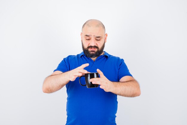 Jeune homme essayant de tenir une tasse avec du thé chaud en chemise bleue et ayant l'air troublé, vue de face.