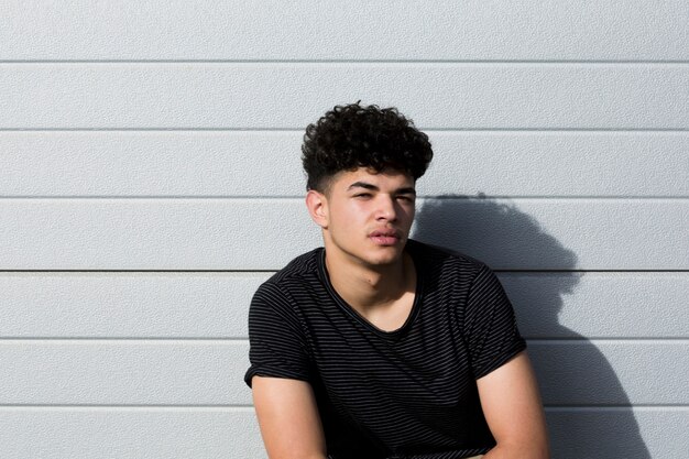 Photo gratuite jeune homme ethnique bouclé en chemise noire assis contre un mur gris