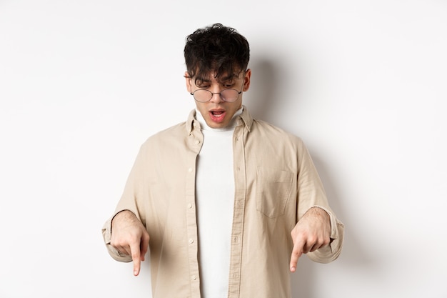 Un jeune homme étonné à lunettes regarde et pointe vers le bas, ouvre la bouche et dit wow impressionné, vérifie l'offre de remise, debout sur fond blanc