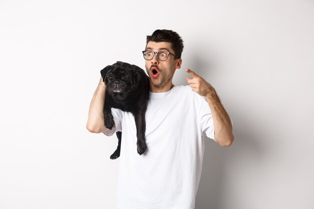 Jeune homme étonné tenant un chien noir mignon sur l'épaule, pointant le doigt vers la gauche de l'offre promotionnelle, regardant impressionné et sans voix, debout sur fond blanc.