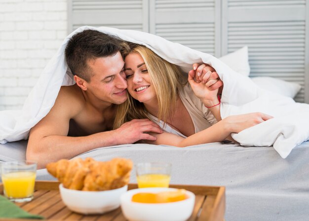 Jeune homme étreignant une femme joyeuse au lit sous une couverture près de la nourriture sur la table du petit déjeuner