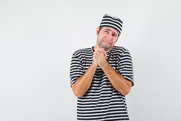 Jeune homme étreignant les mains en geste de prière en pleurant en t-shirt, chapeau et à la recherche d'impuissance. vue de face.