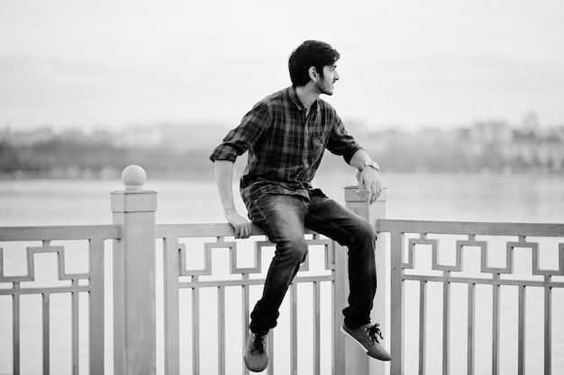 Photo gratuite jeune homme étudiant indien à chemise à carreaux et jeans assis sur des mains courantes contre le lac