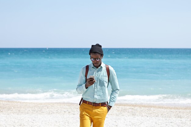 Jeune homme européen occupé à la peau sombre portant des vêtements à la mode et un sac à dos restant en ligne même pendant les vacances, utilisant un téléphone portable sur la plage, ignorant toutes les beautés qui l'entourent