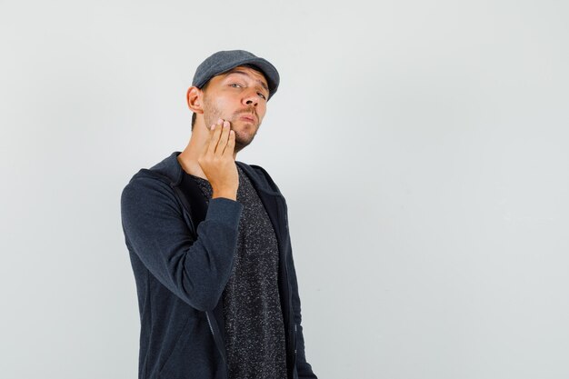 Jeune homme examinant la peau de son visage en t-shirt, veste, casquette et jolie vue de face.