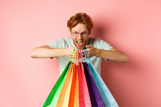 Jeune homme excité, acheteur tenant des sacs à provisions et souriant heureux, l'air excité par les articles achetés, debout sur fond rose