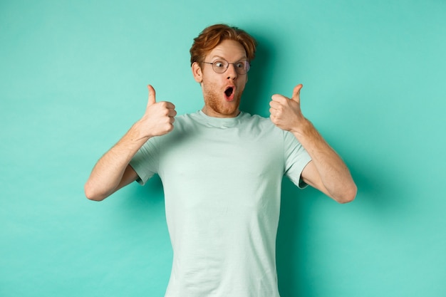 Jeune homme excité aux cheveux rouges et à la barbe, regardant la promo avec admiration, montrant le pouce levé en signe d'approbation, faisant l'éloge de l'offre, debout sur fond turquoise